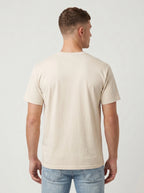 A man viewed from the back wearing a light beige crew-neck t-shirt and light blue jeans, standing against a plain light gray studio background.