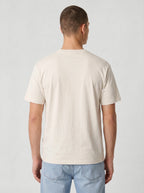 A man viewed from the back wearing a plain cream-colored short-sleeve t-shirt and light blue jeans, standing against a light gray studio background.