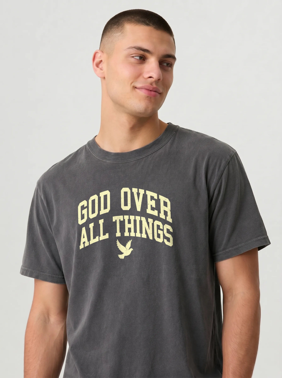 A man wearing a dark gray crewneck t-shirt with 'GOD OVER ALL THINGS' and a dove graphic printed in a light cream color on the chest, set against a plain light gray background.