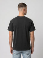 A man viewed from behind wearing a plain black short-sleeve t-shirt and light blue jeans, standing against a neutral light gray background.