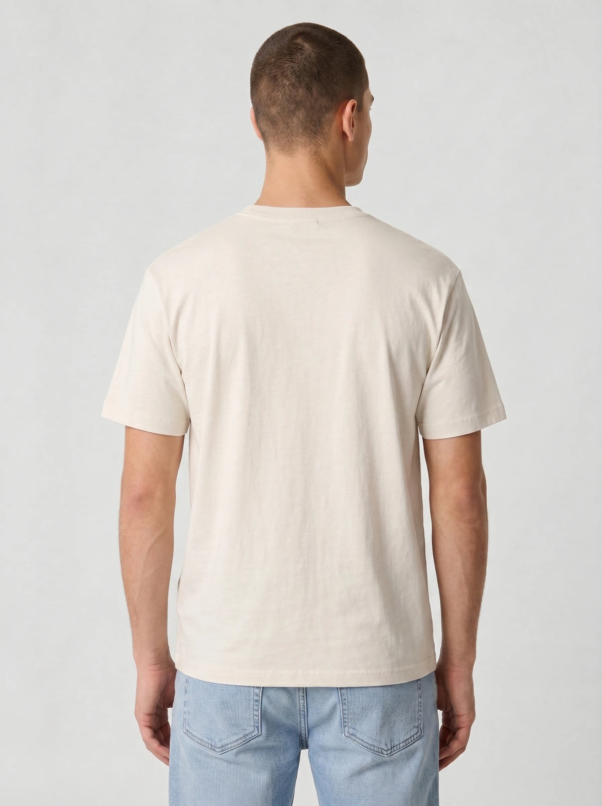 Back view of a man wearing a plain cream Comfort Colors t-shirt with light blue jeans, no graphic on the back, against a light gray studio background.