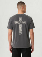 A man viewed from the back wearing a dark gray washed t-shirt with a large distressed cross graphic and white text 'GOD SO LOVED THE WORLD THAT HE GAVE ONLY ME' on the back, paired with black pants, against a light gray studio background.
