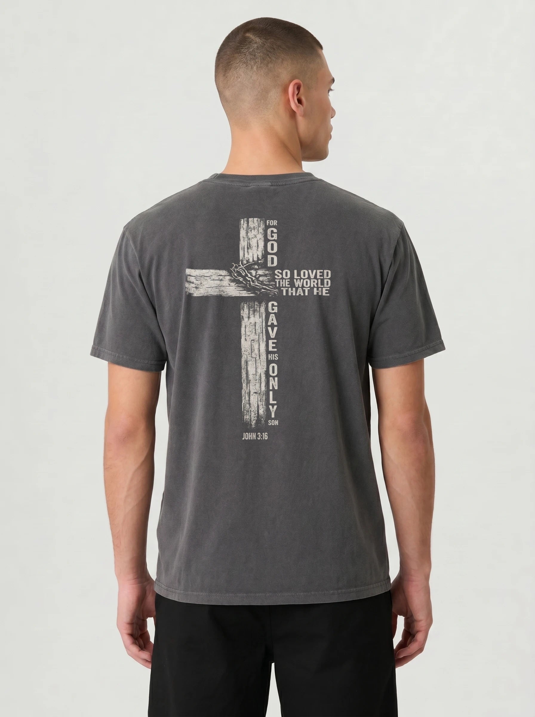 A man viewed from the back wearing a dark gray washed t-shirt with a large distressed cross graphic and white text 'GOD SO LOVED THE WORLD THAT HE GAVE ONLY ME' on the back, paired with black pants, against a light gray studio background.