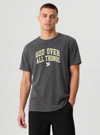A man wearing a dark gray crewneck t-shirt with the text 'GOD OVER ALL THINGS' and a small dove graphic printed in a light beige color on the chest, paired with black pants, against a light gray studio background.