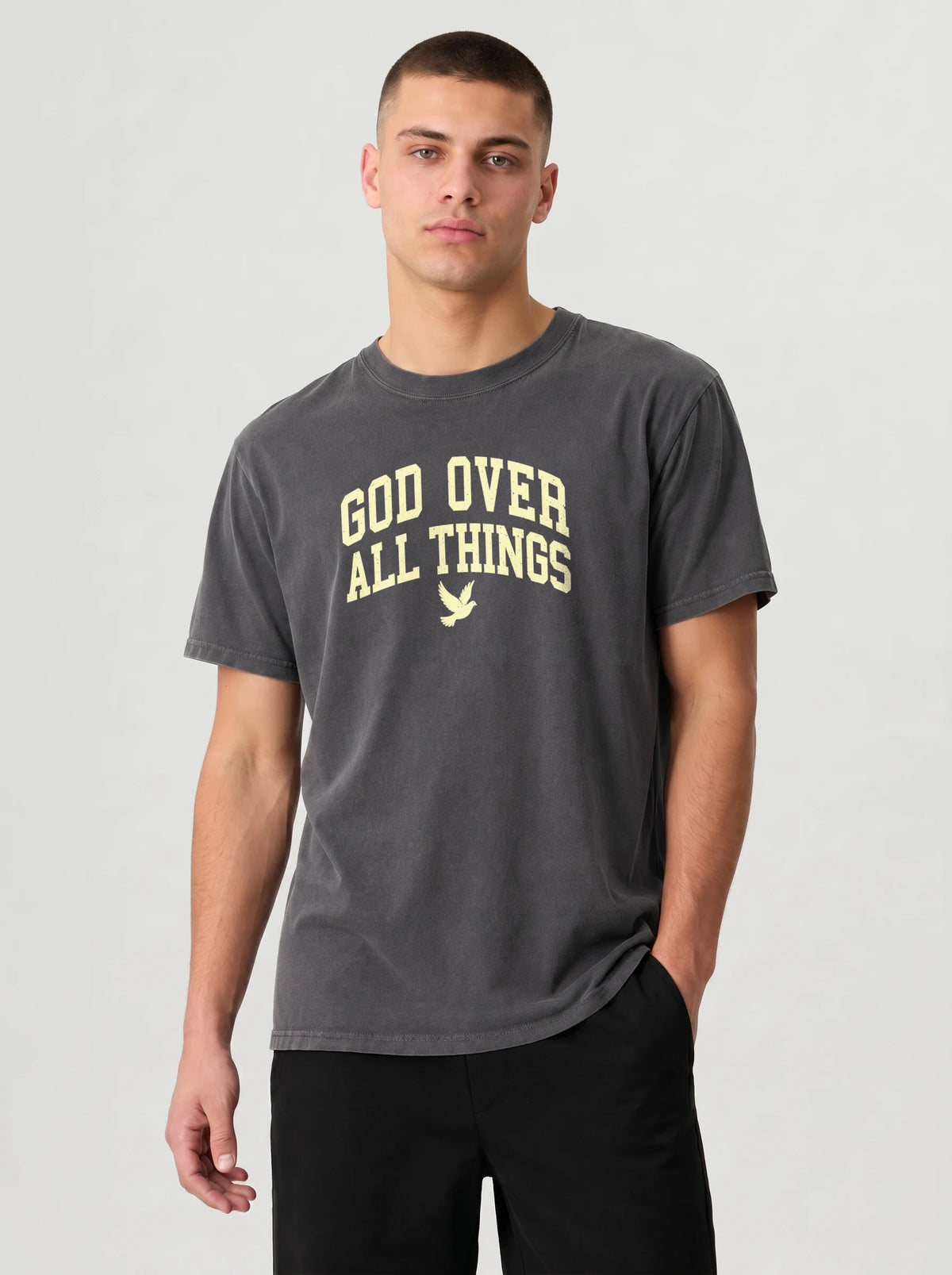 A man wearing a dark gray crewneck t-shirt with the text 'GOD OVER ALL THINGS' and a small dove graphic printed in a light beige color on the chest, paired with black pants, against a light gray studio background.