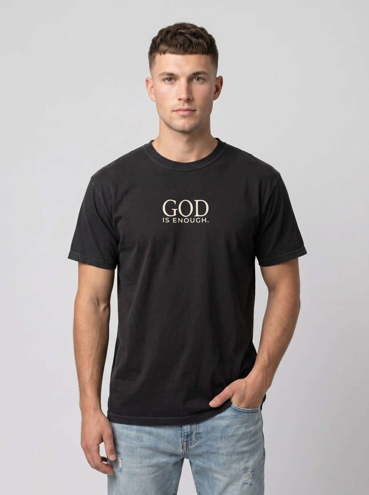 A man wearing a black crew-neck t-shirt with a print that reads 'GOD IS ENOUGH., paired with light blue jeans, against a solid gray studio background.