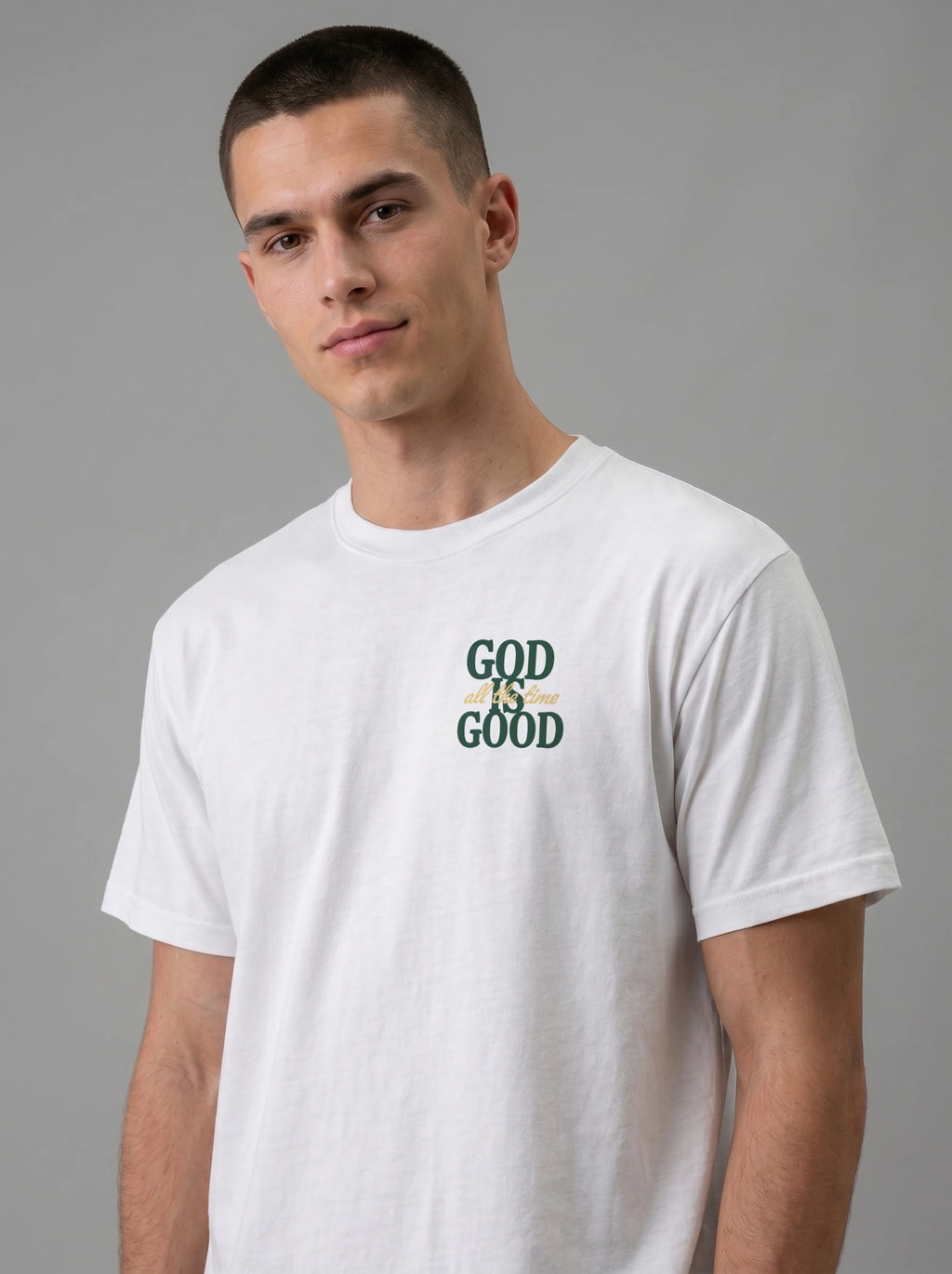 A man wearing a white crew-neck t-shirt with a graphic on the chest that reads 'GOD IS GOOD' in dark green and yellow, set against a solid gray studio background.