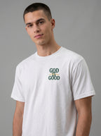 A man wearing a white crew-neck t-shirt with a graphic on the chest that reads 'GOD IS GOOD' in dark green and yellow, set against a solid gray studio background.