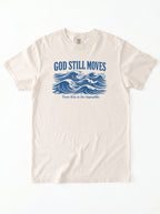 An off-white short-sleeve crewneck t-shirt with a blue graphic print on the front. The graphic features the text 'GOD STILL MOVES' above stylized ocean waves, with the phrase 'Trust Him in the impossible.