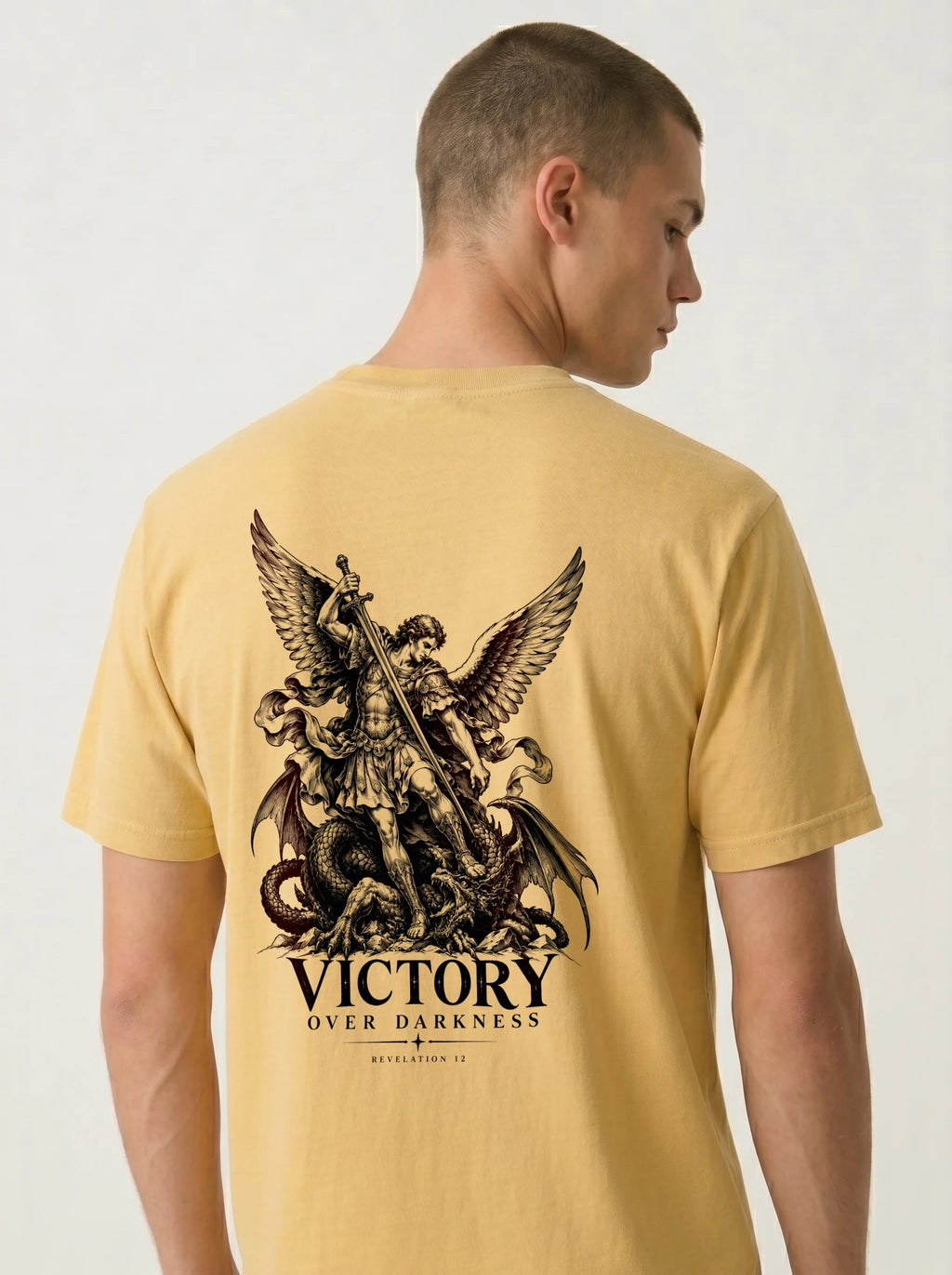 Yellow T-shirt with a winged warrior defeating a dragon and the text “VICTORY OVER DARKNESS” and “REVELATION 12.”