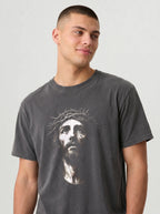 A man wearing a dark gray crewneck t-shirt with a white graphic print of Jesus' face wearing a crown of thorns, photographed against a light gray studio background.