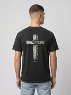 A man viewed from the back wearing a black t-shirt with a large white graphic of a cross and biblical text 'GOD SO LOVED THE WORLD THAT HE GAVE ONLY ME', paired with light blue jeans, against a plain light gray background.