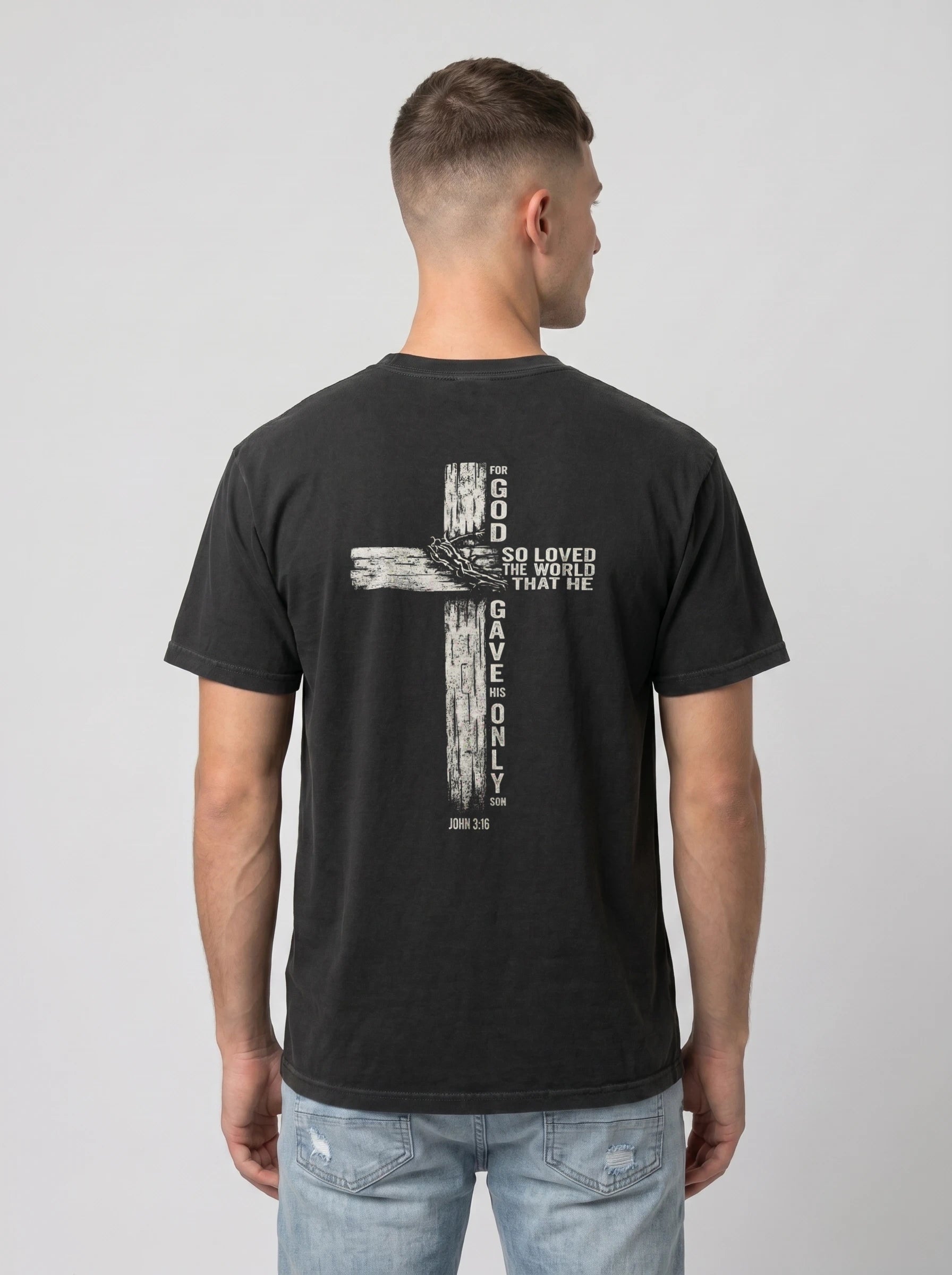 A man viewed from the back wearing a black t-shirt with a large white graphic of a cross and biblical text 'GOD SO LOVED THE WORLD THAT HE GAVE ONLY ME', paired with light blue jeans, against a plain light gray background.