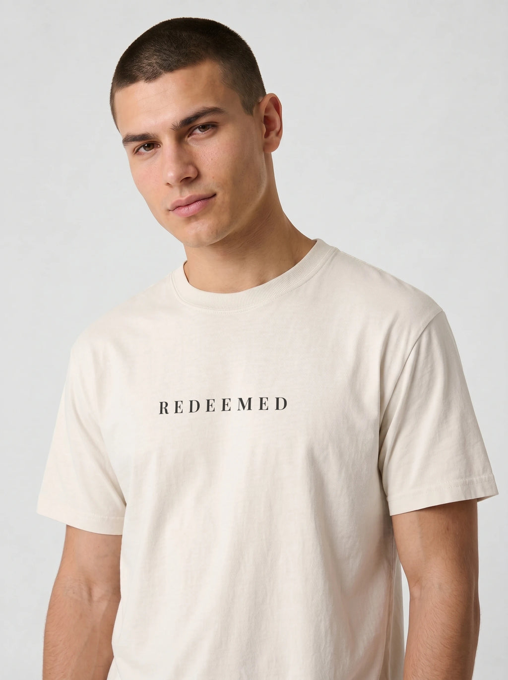 A man wearing a light beige crewneck t-shirt with the word 'REDEEMED' printed in black serif letters across the chest, photographed against a neutral light gray background.