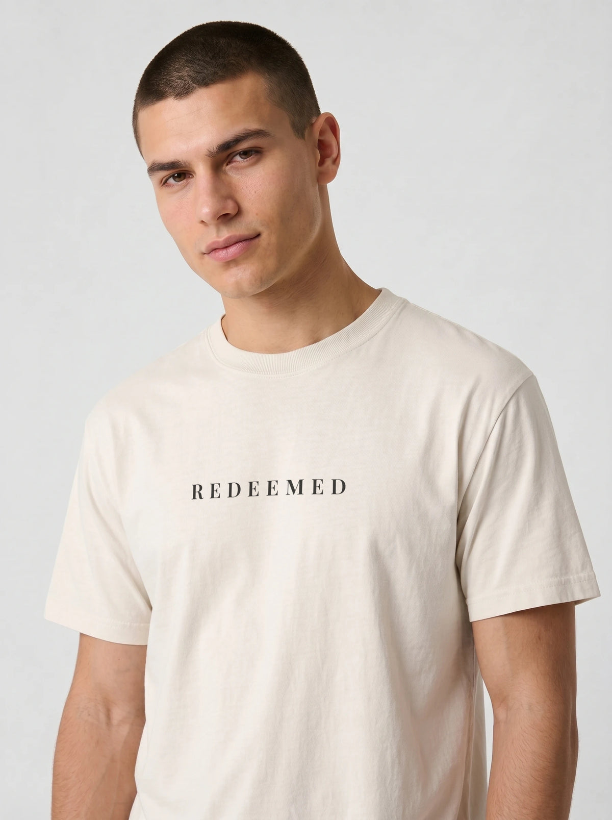 A man wearing a light beige crewneck t-shirt with the word 'REDEEMED' printed in black serif letters across the chest, photographed against a neutral light gray background.
