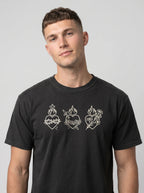 A man wearing a black crewneck t-shirt with three white Sacred Heart of Jesus line-art graphics across the chest, photographed against a neutral gray background.