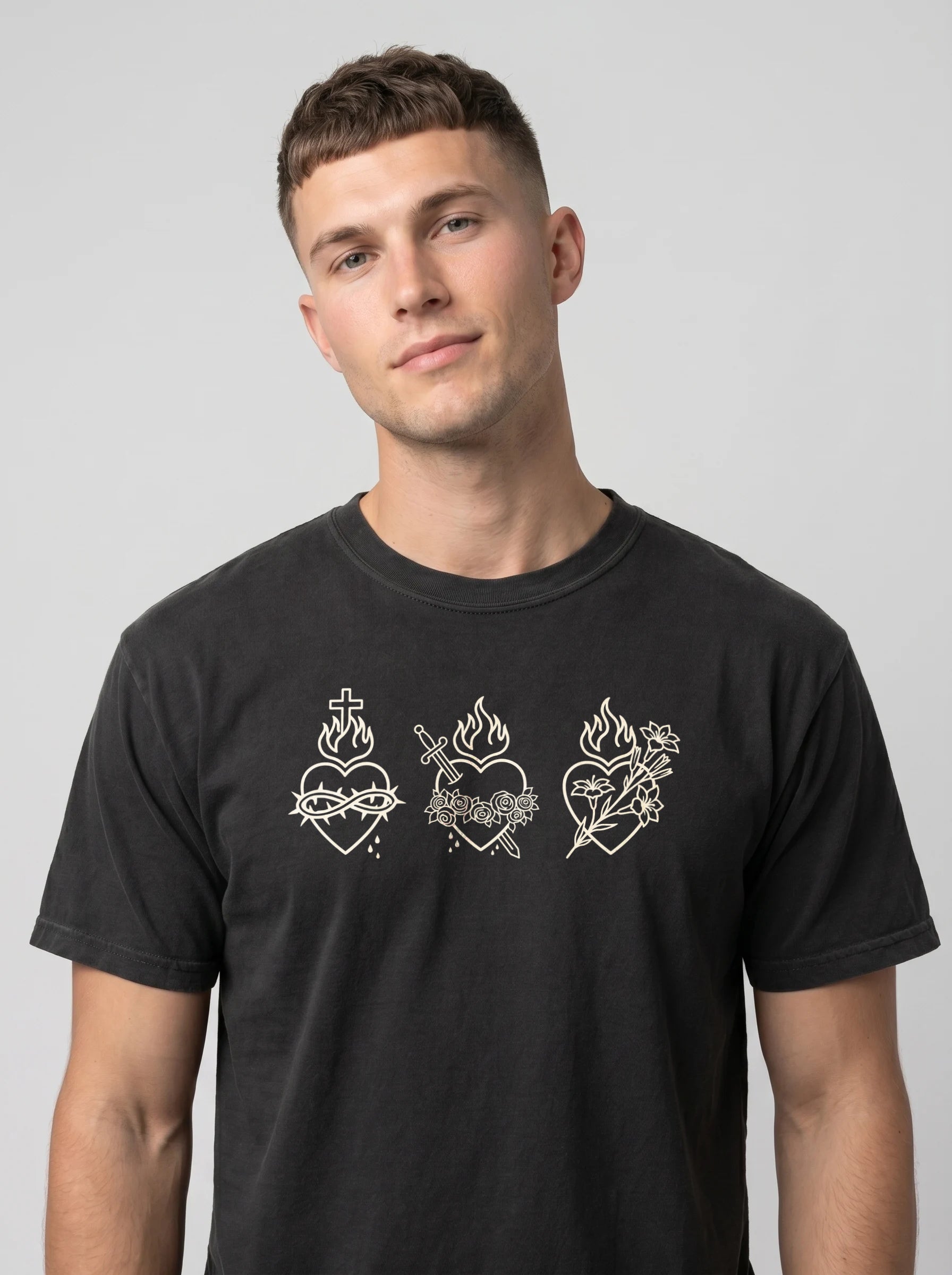 A man wearing a black crewneck t-shirt with three white Sacred Heart of Jesus line-art graphics across the chest, photographed against a neutral gray background.