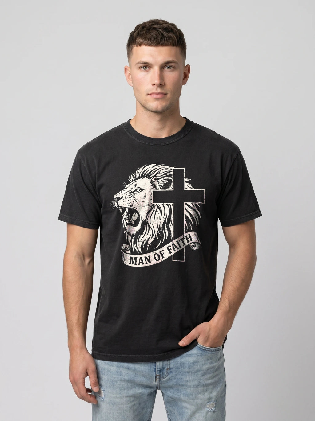 A man wearing a black crewneck t-shirt with a white graphic of a lion and cross, with the text 'MAN OF FAITH' printed on the front, paired with light blue jeans, against a solid gray studio background.