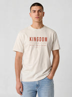 A man wearing a cream-colored t-shirt with 'KINGDOM NOT CULTURE' printed in red on the chest, paired with light blue jeans, against a light gray studio background.