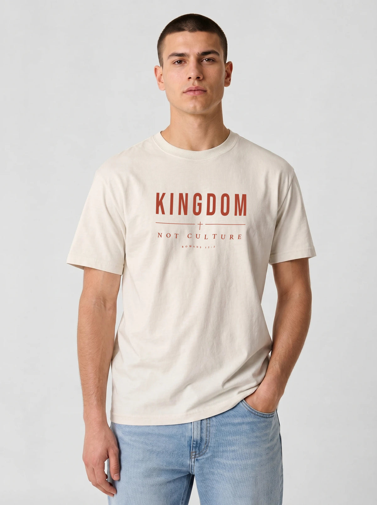 A man wearing a cream-colored t-shirt with 'KINGDOM NOT CULTURE' printed in red on the chest, paired with light blue jeans, against a light gray studio background.
