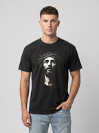 A man wearing a black crew-neck t-shirt with a high-contrast graphic print of Jesus' face on the front, paired with light-wash blue jeans, against a solid gray studio background.