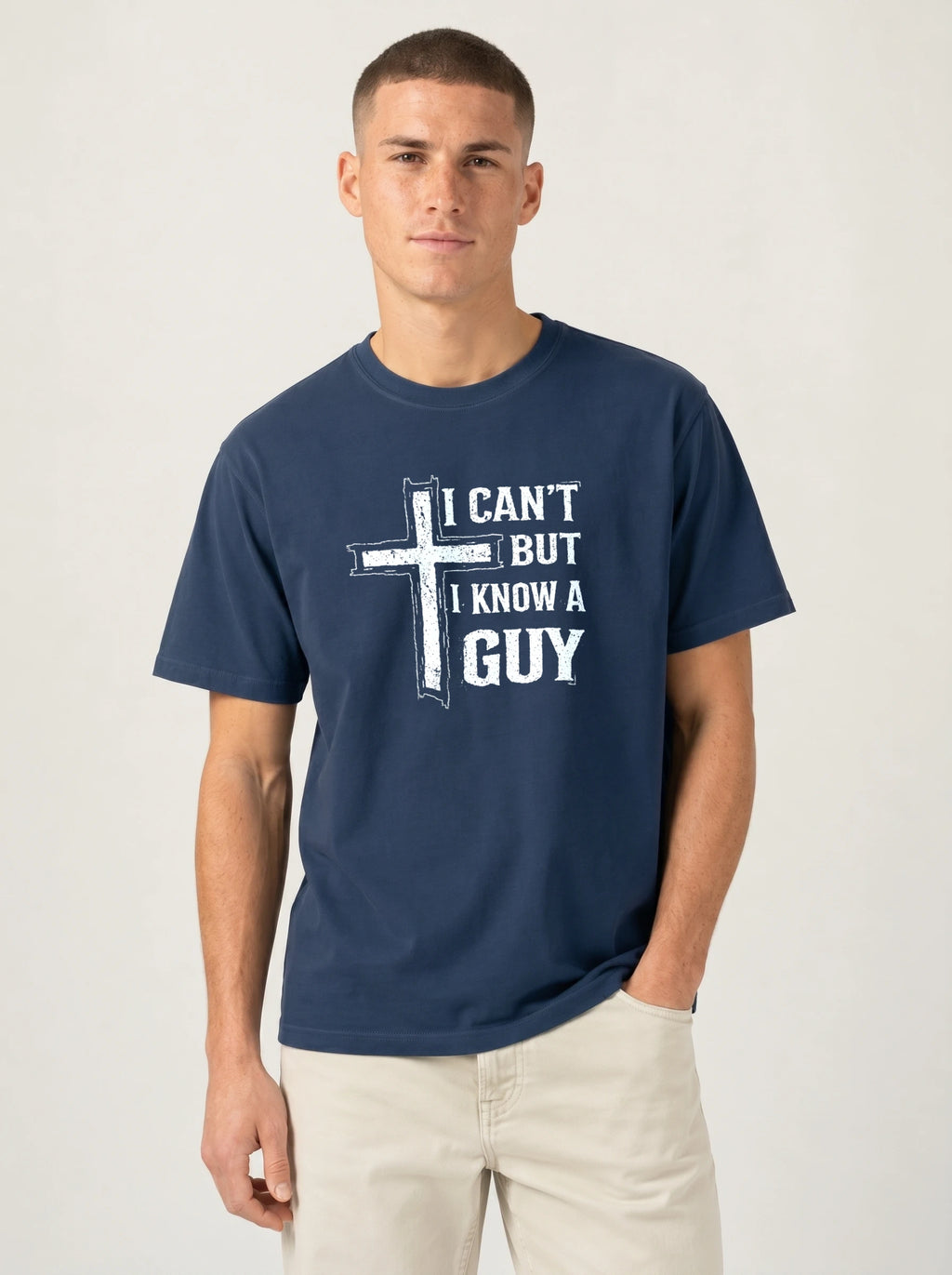 A man wearing a navy blue t-shirt with a white graphic print that reads 'I CAN'T BUT I KNOW A GUY' alongside a cross design, paired with light beige pants, against a plain light gray background.