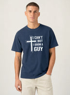 A man wearing a navy blue t-shirt with a white graphic print that reads 'I CAN'T BUT I KNOW A GUY' alongside a cross design, paired with light beige pants, against a plain light gray background.