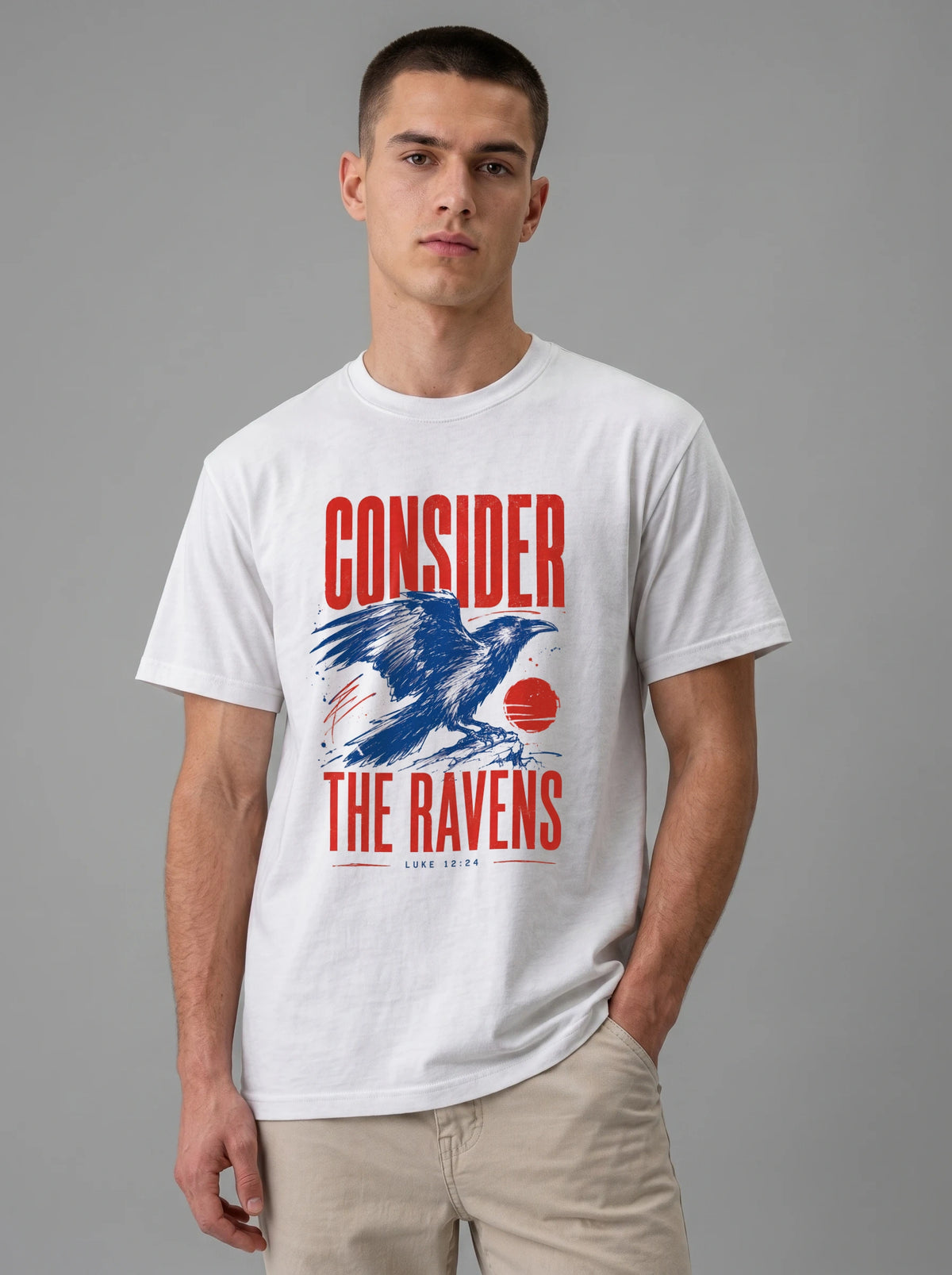 A man wearing a white graphic t-shirt with a red and blue raven design and the text 'CONSIDER THE RAVENS', paired with beige pants, against a solid gray studio background.