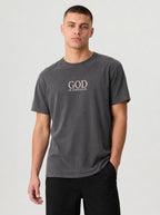 A man wearing a dark gray crewneck t-shirt with the text 'GOD IS ENOUGH.' printed in a light beige serif font on the chest, paired with black pants, against a light gray studio background.