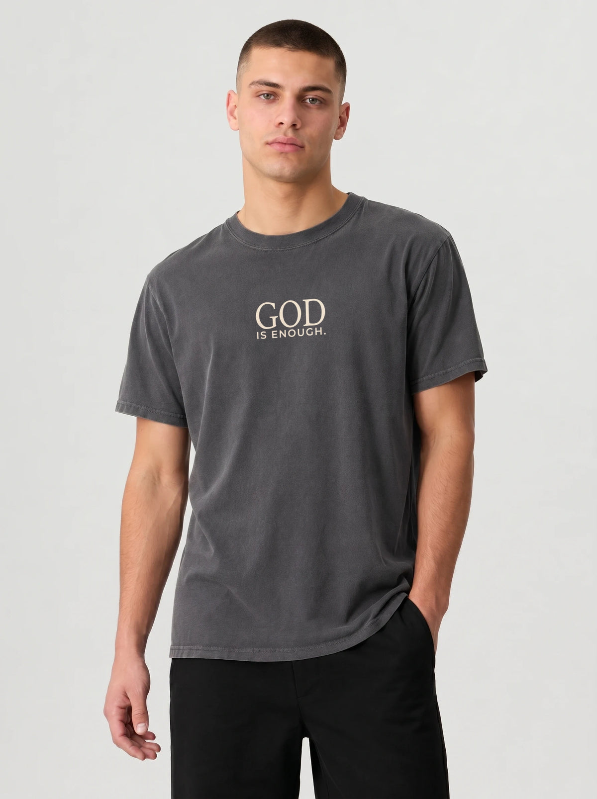 A man wearing a dark gray crewneck t-shirt with the text 'GOD IS ENOUGH.' printed in a light beige serif font on the chest, paired with black pants, against a light gray studio background.