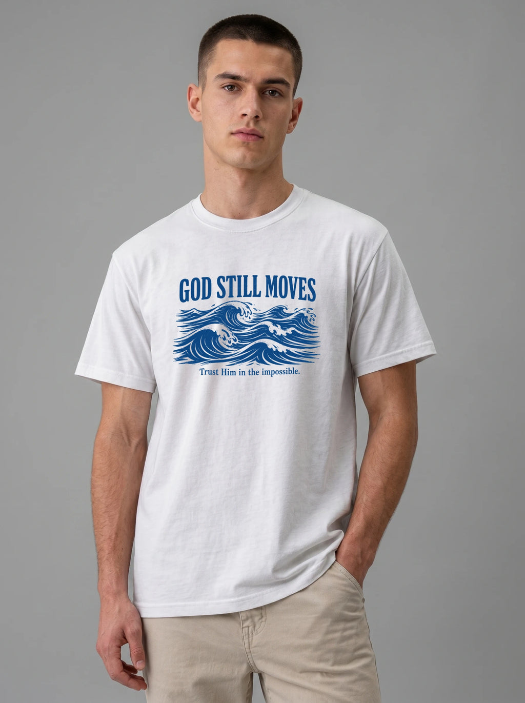 A man wearing a white crewneck t-shirt with a blue graphic print reading "GOD STILL MOVES" above ocean waves and "Trust Him in the impossible," paired with khaki pants against a solid gray studio background.