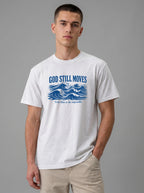 A man wearing a white crewneck t-shirt with a blue graphic print reading "GOD STILL MOVES" above ocean waves and "Trust Him in the impossible," paired with khaki pants against a solid gray studio background.