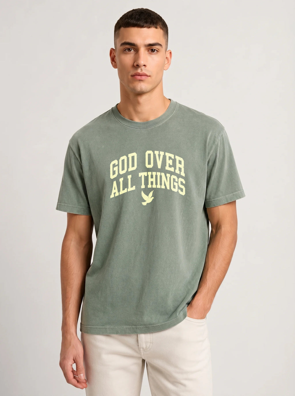 A man wearing a sage green crewneck t-shirt with 'GOD OVER ALL THINGS' printed in cream on the chest, paired with light beige pants, against a light gray studio background.