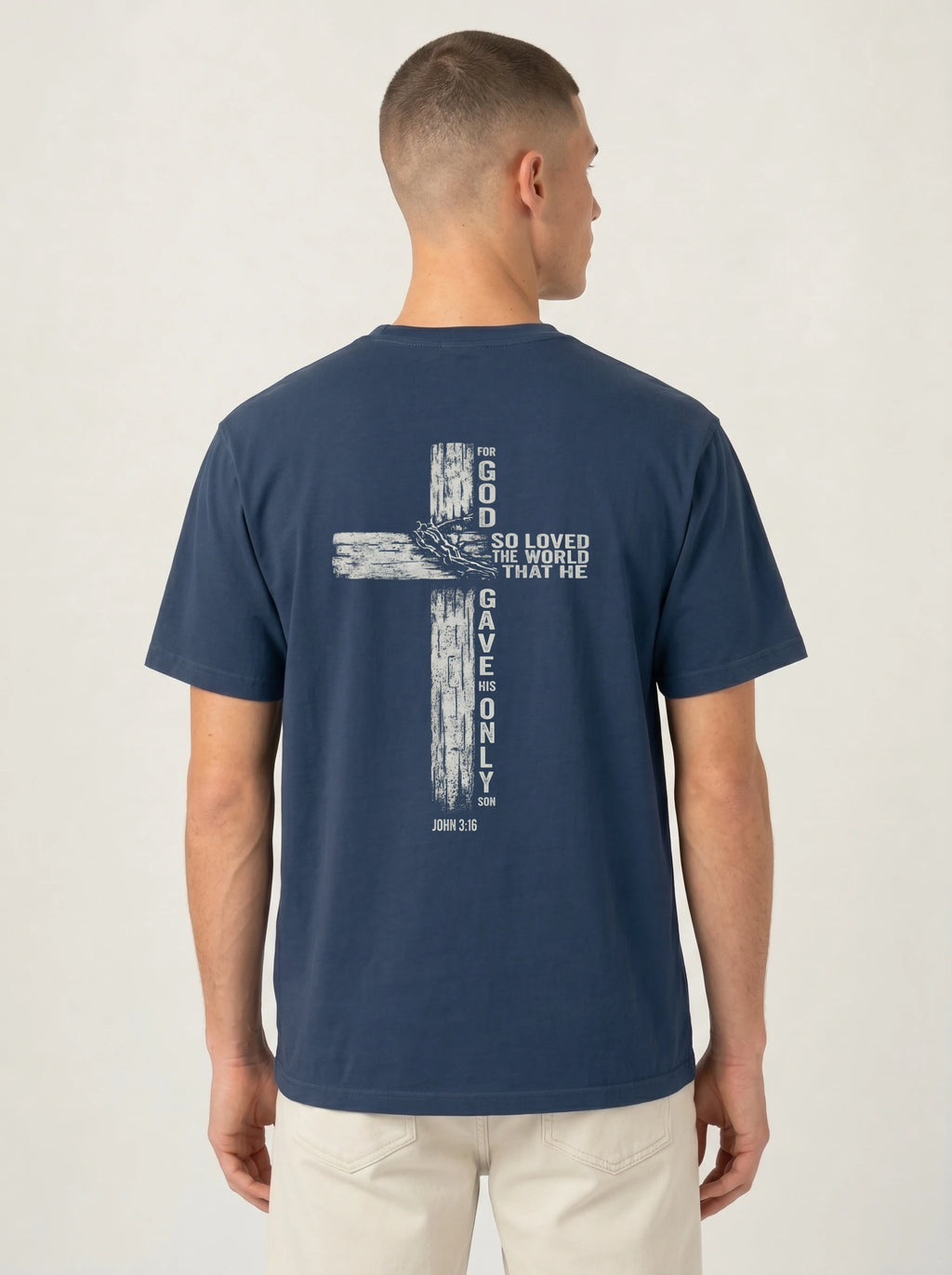 A man viewed from the back wearing a navy blue t-shirt with a large white distressed cross graphic and the text 'GOD SO LOVED THE WORLD THAT HE GAVE ONLY ONE' printed on the back, paired with light beige pants against a light gray studio background.