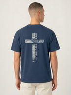 A man viewed from the back wearing a navy blue t-shirt with a large white distressed cross graphic and the text 'GOD SO LOVED THE WORLD THAT HE GAVE ONLY ONE' printed on the back, paired with light beige pants against a light gray studio background.