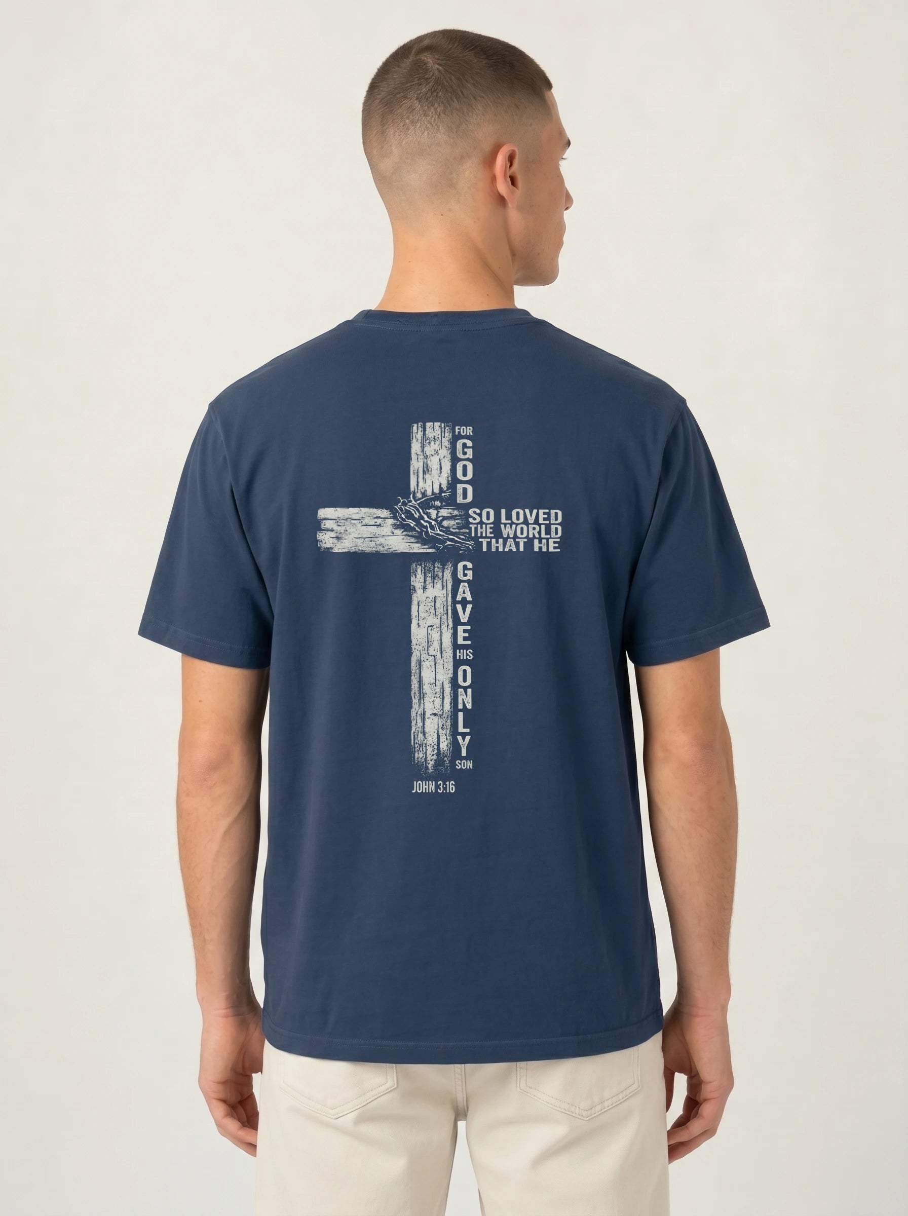 A man viewed from the back wearing a navy blue t-shirt with a large white distressed cross graphic and the text 'GOD SO LOVED THE WORLD THAT HE GAVE ONLY ONE' printed on the back, paired with light beige pants against a light gray studio background.