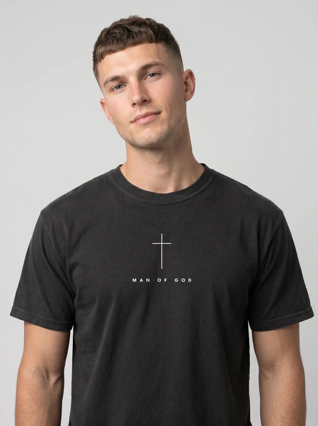 A man wearing a black crewneck t-shirt with the text 'MAN OF GOD' printed in white letters on the chest, photographed against a solid light gray background.