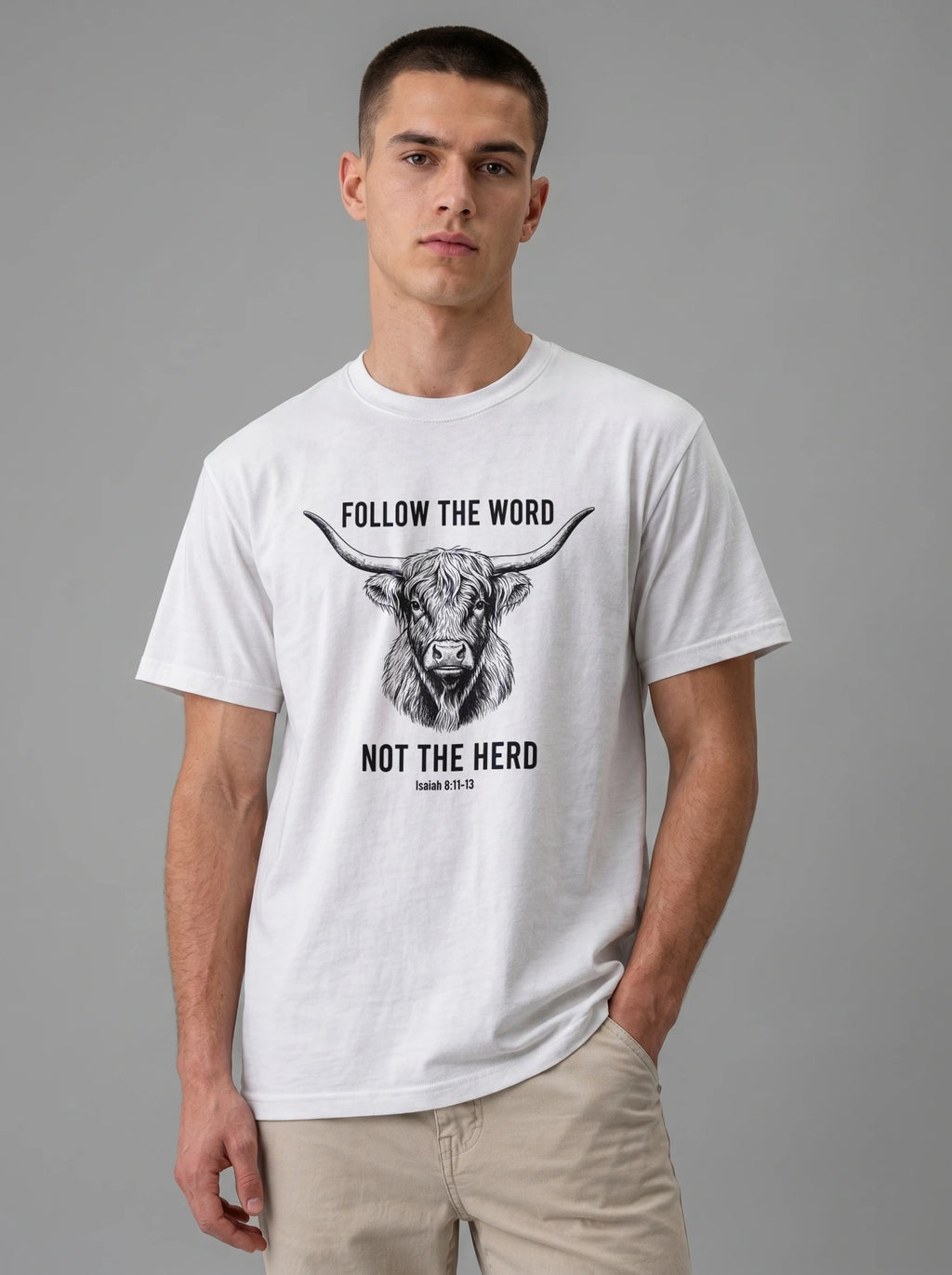 A man wearing a white short-sleeve graphic t-shirt with a black illustration of a longhorn cow and the text 'FOLLOW THE WORD NOT THE HERD' on the front, paired with beige pants, against a solid gray studio background.