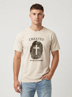 A man wearing a light beige crewneck t-shirt with a black graphic print of a fingerprint containing a cross and the words 'CREATED WITH A PURPOSE'.