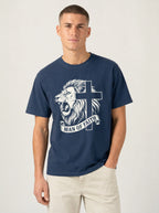 A man wearing a navy blue crewneck t-shirt with a white graphic of a lion and cross, with the text 'MAN OF FAITH' printed below, paired with light beige pants, against a plain light gray background.