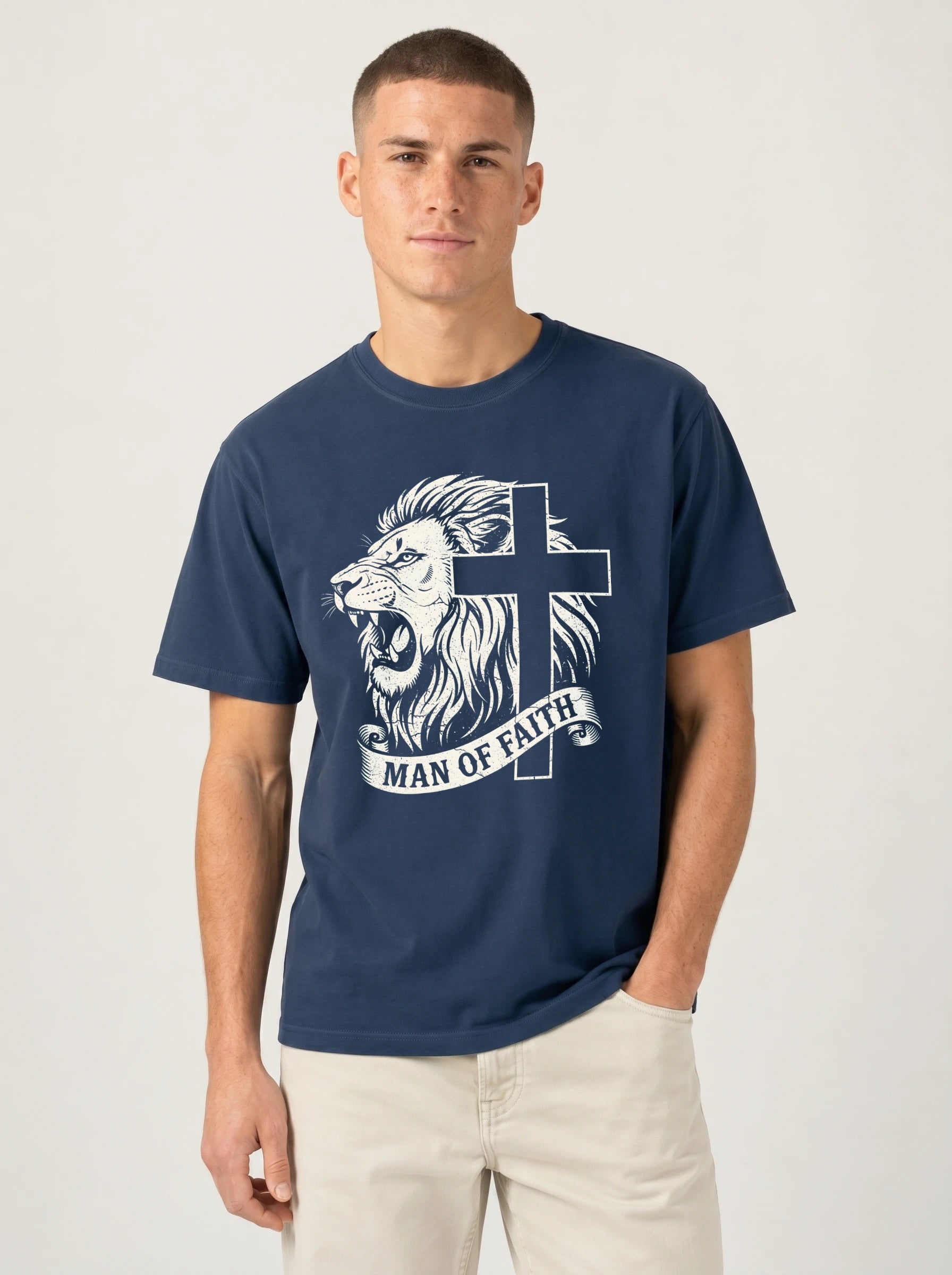 A man wearing a navy blue crewneck t-shirt with a white graphic of a lion and cross, with the text 'MAN OF FAITH' printed below, paired with light beige pants, against a plain light gray background.