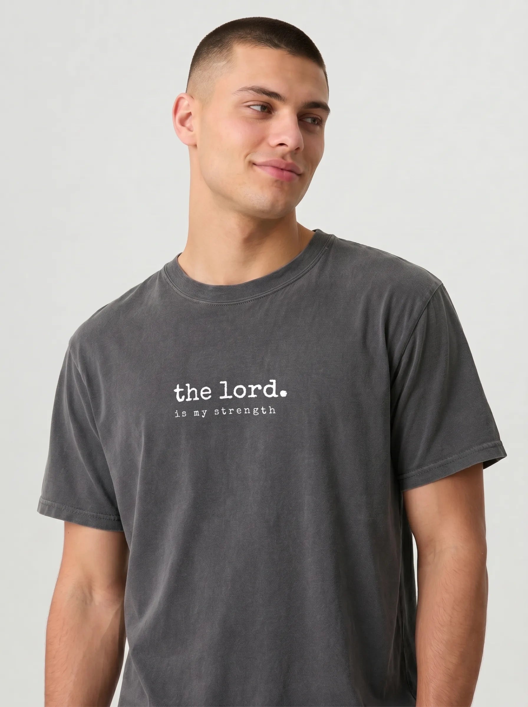 A man wearing a dark gray washed t-shirt with the white printed text 'the lord. is my strength' on the chest, photographed against a light gray background.