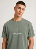 Person wearing a muted green T-shirt with a white cross above the text “Set Apart” and “Romans 1:1.”