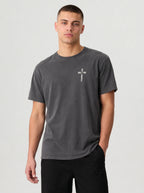 A man wearing a dark gray crewneck t-shirt with a small white cross graphic on the left chest, paired with black pants, against a light gray studio background.