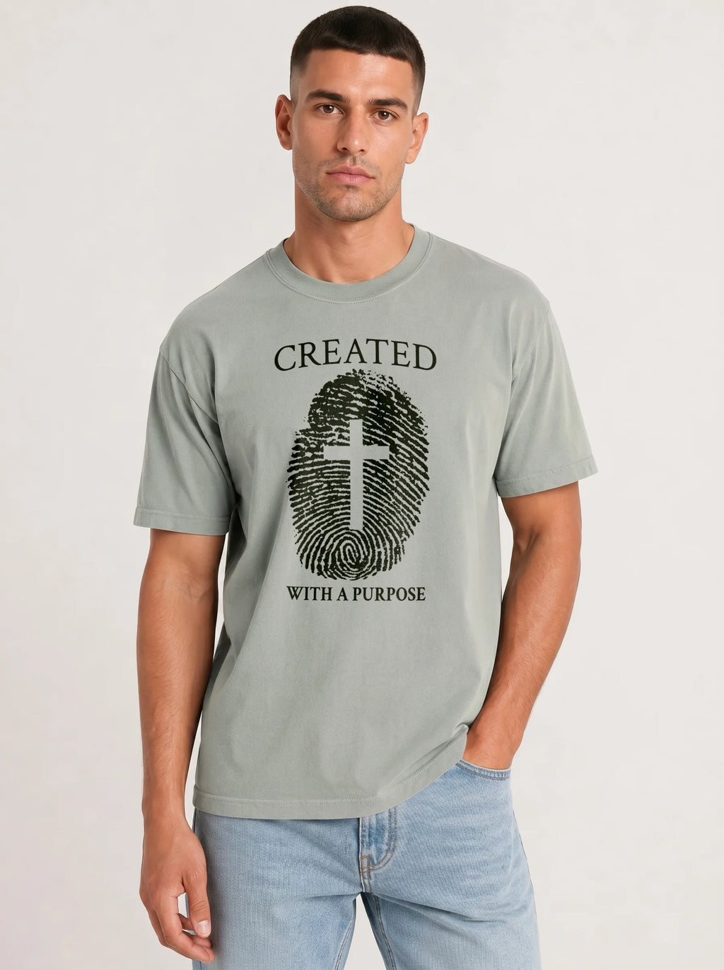 A man wearing a light gray crewneck t-shirt with a black graphic print of a fingerprint and cross with the text 'CREATED WITH A PURPOSE'.