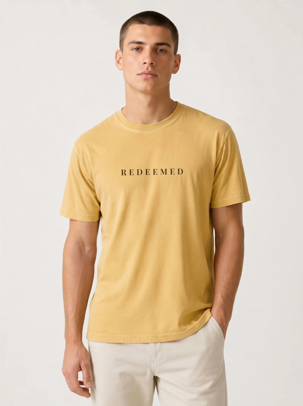 A man wearing a mustard yellow crewneck t-shirt with the word 'REDEEMED' printed in black across the chest, paired with off-white pants, against a plain light gray background.