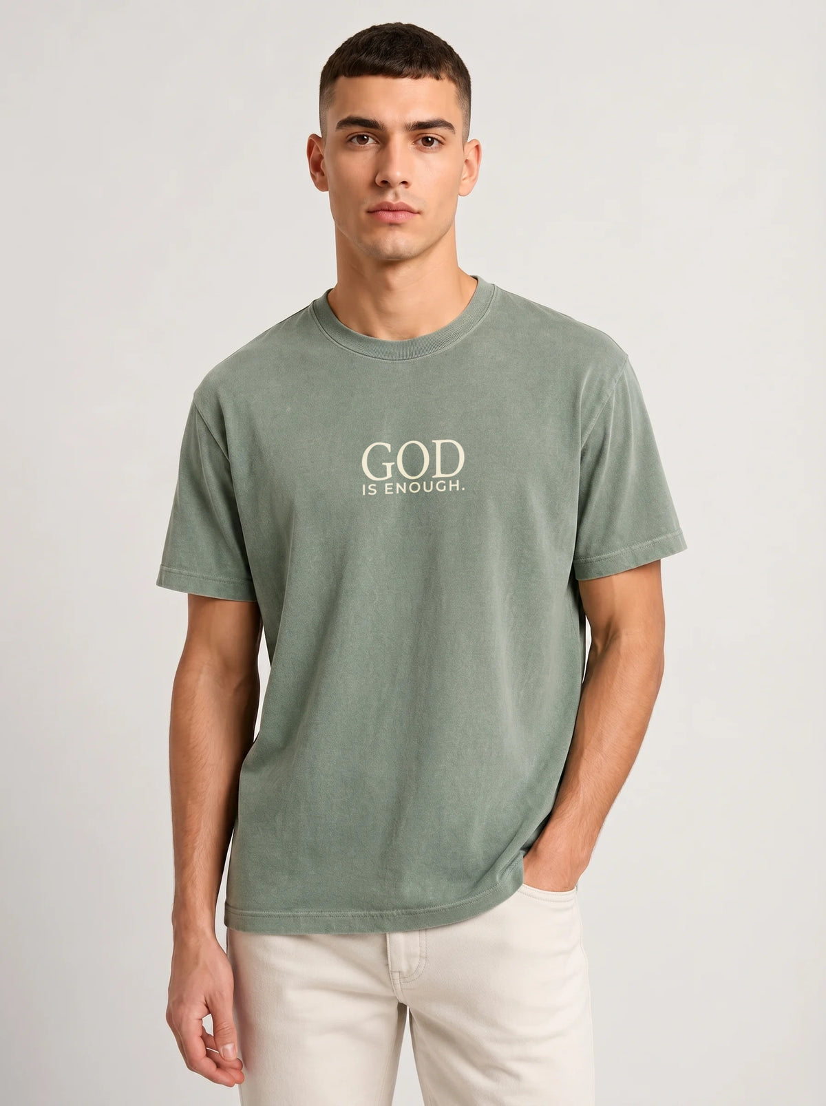 A man wearing a sage green crewneck t-shirt with the text 'GOD IS ENOUGH.' printed in cream on the chest, paired with off-white pants, against a light gray studio background.