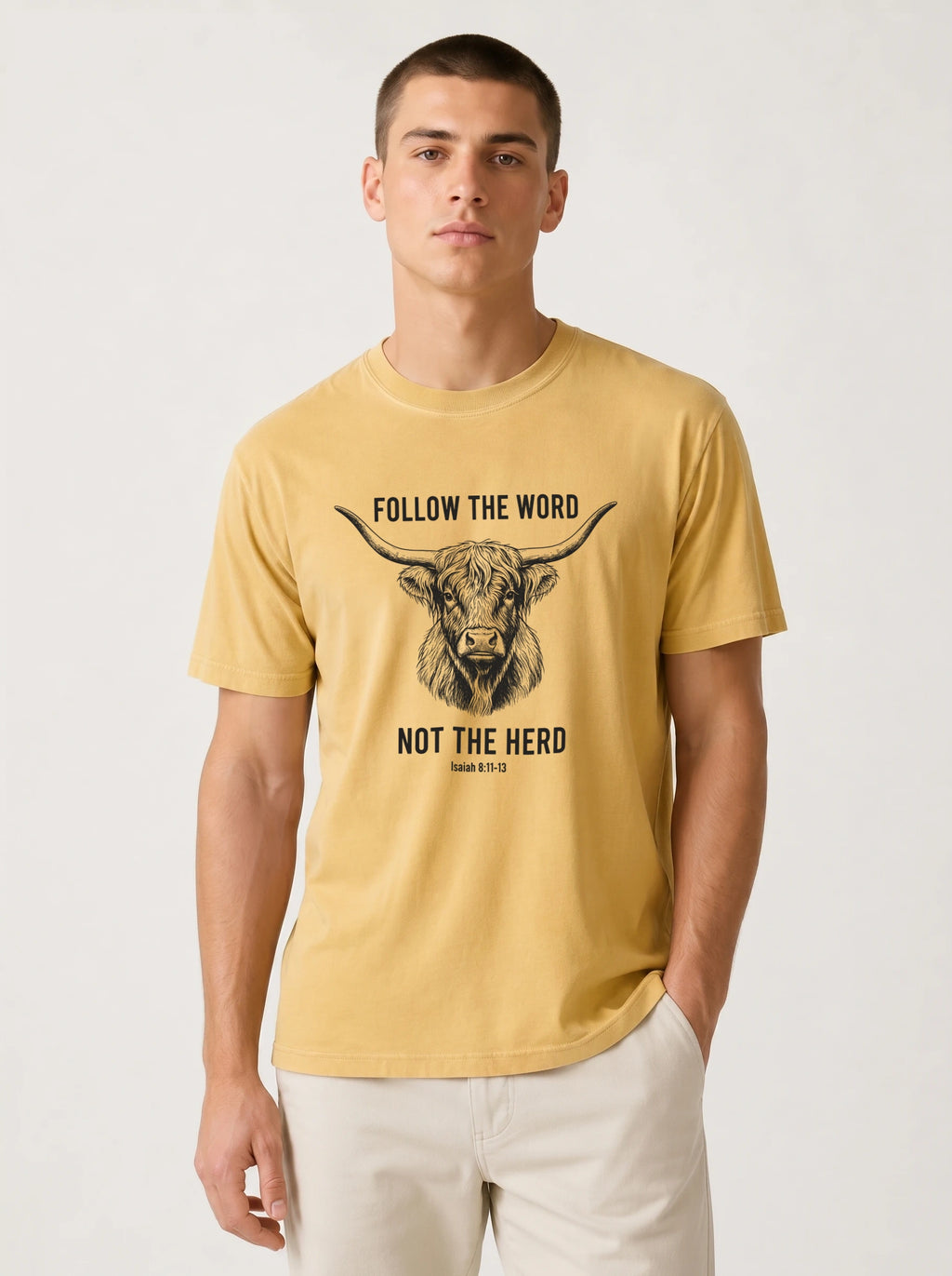 A man wearing a mustard yellow graphic t-shirt with a black bull illustration and the text 'FOLLOW THE WORD NOT THE HERD' on the front, paired with off-white trousers, against a plain light gray background.