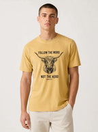 A man wearing a mustard yellow graphic t-shirt with a black bull illustration and the text 'FOLLOW THE WORD NOT THE HERD' on the front, paired with off-white trousers, against a plain light gray background.