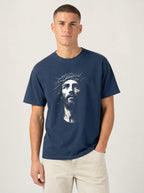 A man wearing a navy blue crewneck t-shirt with a graphic print of Jesus Christ's face on the front, paired with light beige pants, against a plain light gray background.